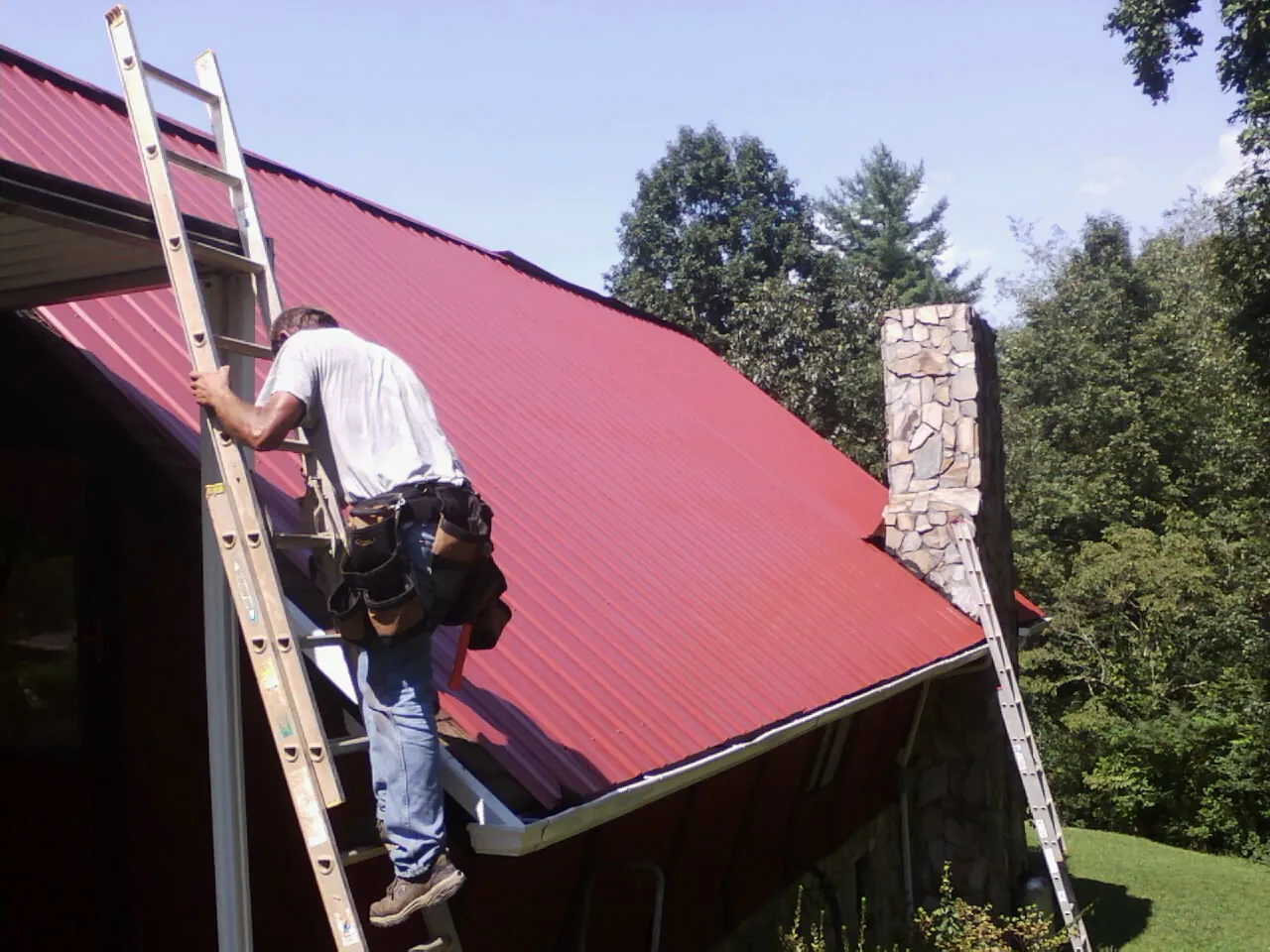 Roofer on a ladder beside a red metal roof and stone chimney — Soffit & Fascia Repair by Roofing Brothers in Hoskins